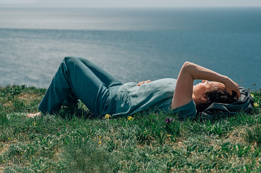 an adult woman, a pensioner, lies on the grass barefoot with a smile on her face on a sunny day on a mountain against the backdrop of the sea in summer, spring. High quality photo