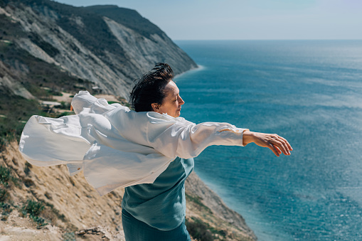 adult woman with outstretched arms, like wings, stands on top of a mountain against the backdrop of the sea. a person is freed from worries, begins a new life, with pleasure exposes his face to the sun and wind. High quality photo