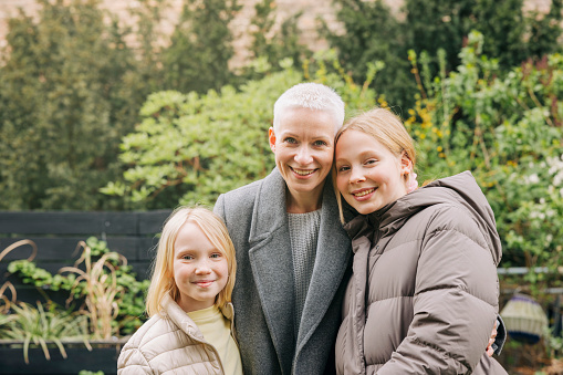 Portrait of mother with her two daughters standing together in backyard garden. Woman with her two beautiful girls in warm clothing looking at camera and smiling.