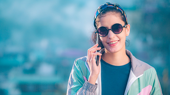 In this image with copy space, an Asian/Indian beautiful, happy young woman uses a smartphone in the hills of Himachal Pradesh. She is in a jacket and dark sunglasses.