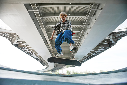 Graphic action shot of young teenager riding skateboard in urban area and jumping in air doing tricks under concrete bridge structure, copy space