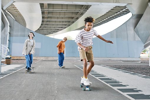 Full length portrait of young teenage boy riding skateboard with friends at skatepark outdoors, copy space