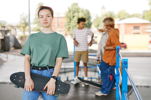Waist up portrait of teenage girl with skateboard looking at camera diverse group of friends in background, copy space