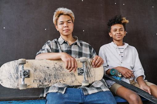 Portrait of two active teenage boys with skateboards sitting on ramp and smiling at camera