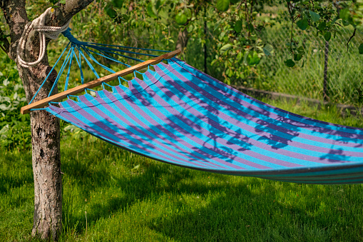 Striped blue and blue fabric hammock hanging in the shade of trees in the garden on a sunny warm day. Relaxation and passive rest from the hustle and bustle of the city