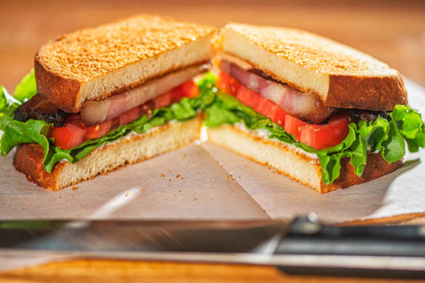 A sliced Bacon, lettuce and tomato on white bread toasted with mayonnaise sitting on a piece of wax paper on a wooden cutting board with knife