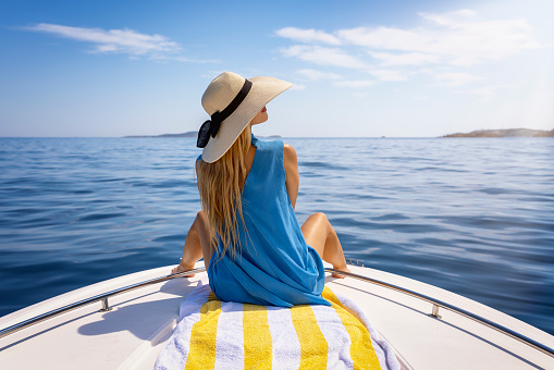 A beautiful, blonde woman with a hat sits on a boat and enjoys the calm sea during her summer holidays