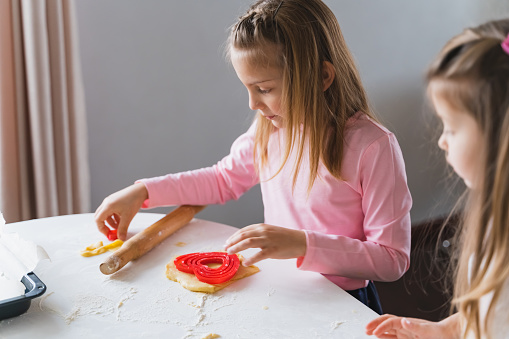 Little girls, sisters cooking homemade heart shaped cookies for valentine's day. Holiday for all lovers. Gift, surprize for mom. Red molds, rolling pin, flour, dough.Handmade family, bakery with kids.