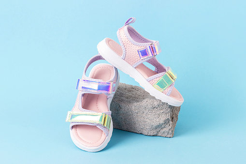 Stylish holographic sandals for kids on grey stone, blue background. Shiny fashion summer shoes. Front view.