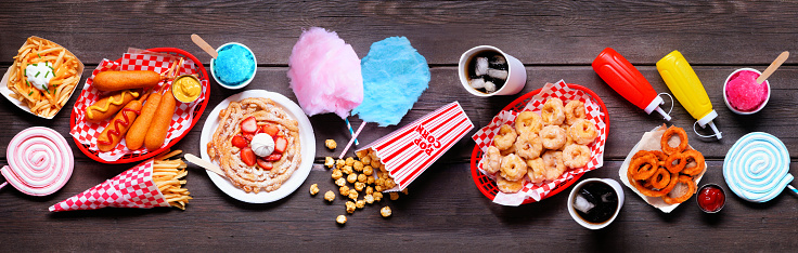 Carnival theme food table scene over a dark wood banner background. Above view. Summer fair concept. Corn dogs, funnel cake, cotton candy and snacks.