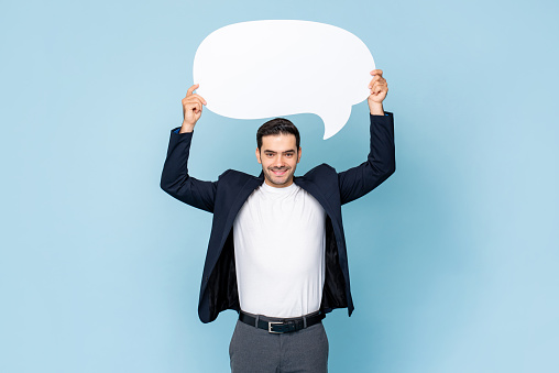 Portrait of young handsome Caucasian man in formal dress holding speech bubble on isolated light blue studio background