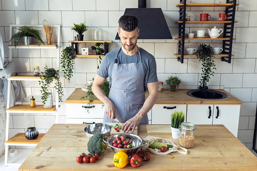 Handsome father, strong young man cooking healthy vegetable salad with fresh organic ingredients, tasty food in the kitchen at home . Men doing chores. Ripe pepper, tomato, cucumber