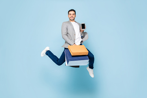Young smiling Caucasian man holding shopping bags and mobile phone while jumping in light blue isolated background