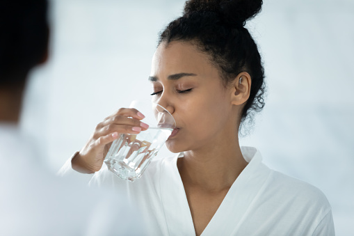Mirror reflection happy peaceful beautiful young african american woman in bathrobe drinking glass of fresh pure aqua, enjoying daily healthcare habit, sipping warm water preventing dehydration.