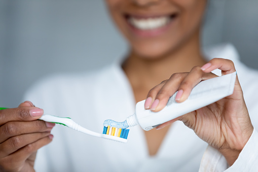 Close up young smiling african american woman applying whitening paste on toothbrush, doing toothcare procedures at home, taking care of gums health, preventing caries, healthy daily habit concept.
