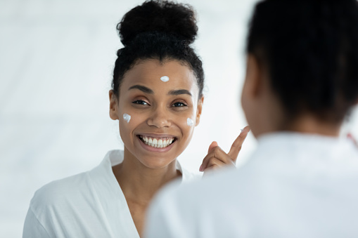 Mirror reflection attractive young african american woman in bathrobe applying moisturizing smoothing cream on face, excited by effective cosmetics results, enjoying morning skincare beauty routine.