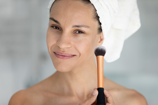 Head shot portrait of happy beautiful hispanic latina woman with towel on head holding clean brush in hands, starting doing daily makeup using tool, applying foundation getting ready in morning.