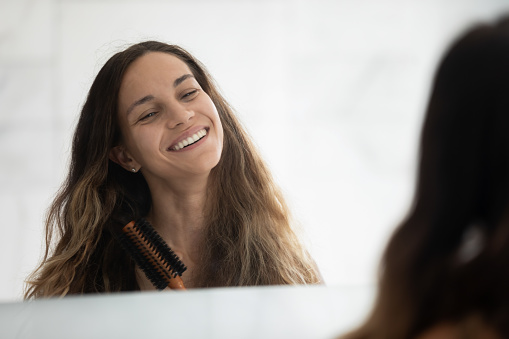 Head shot mirror reflection happy young attractive mixed race woman holding round brush, styling hair in bathroom. Joyful hispanic lady combing long hair, doing hairstyle after morning shower at home.