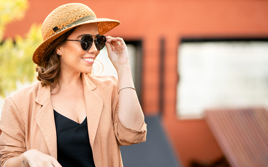 Young woman wearing a sun hat and sunglasses smiling while sitting outside on a rooftop terrace in the summertime