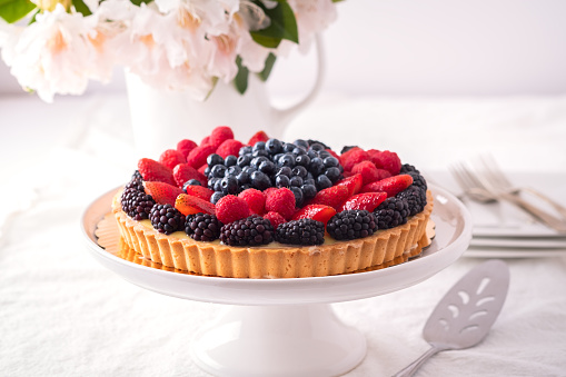 Patriotic celebration - fresh blackberry, strawberry, raspberry and blueberry flan, silverware, white flowers, cake stand, plates, and table cloth