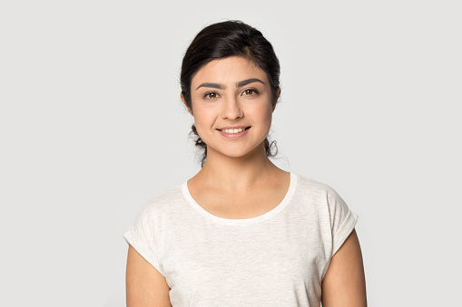 Profile picture of smiling millennial Indian woman isolated on gray studio wall background feeling confident. Headshot portrait of happy young mixed race female intern employee. Recruitment concept.
