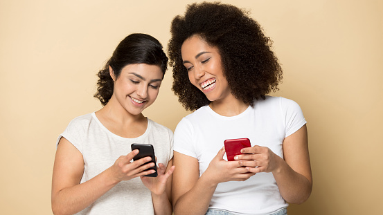 Happy millennial diverse girlfriends isolated on yellow studio background use modern smartphone gadgets chatting online. Smiling young multiracial female friends text message on cellphone devices.