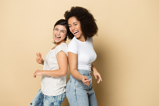 Overjoyed young multiethnic girlfriends isolated on yellow studio background dance have fun together. Smiling happy millennial diverse female friends laugh entertain. Diversity, humor concept.