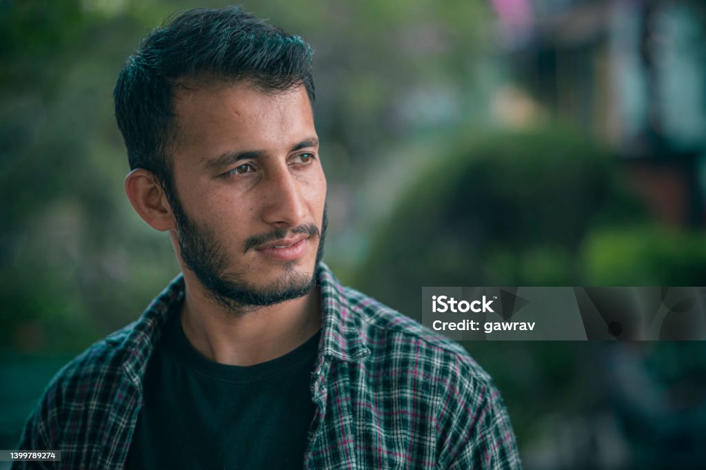 Outdoor day portrait of a young man contemplates and looks away. An Asian/Indian handsome young man contemplates looking at a view with a blank expression in this outdoor day image with copy space and selective focus on the foreground. He stands in a park wearing a casual dress. Adult Stock Photo