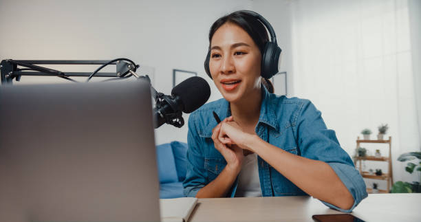 Happy asia girl record a podcast on laptop computer with headphones and microphone talk with audience at living room. Happy asia girl record a podcast on laptop computer with headphones and microphone talk with audience at living room. Female podcaster make audio podcast from her home studio. Stay at house concept. web radio stock pictures, royalty-free photos & images