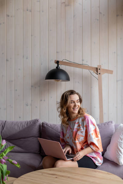 la charmante femme aux cheveux bruns riante avec les cheveux longs dans des vêtements confortables travaille à la maison sur internet assise sur un canapé confortable contre un mur en bois. . intérieur moderne. travaillez à la maison en ligne. - women computer home interior brown hair photos et images de collection