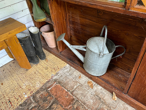 Stock photo showing inside a wooden shed stood on brick floor, storing a metal watering can, a flower pot, and pairs of green and black wellington boots.