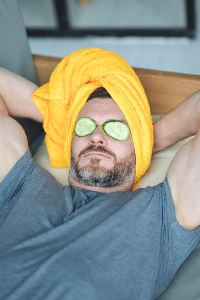 hombre guapo acostado en el sofá con rodajas de pepino en los ojos y toalla amarilla en la cabeza. concepto de autocuidado masculino, cuidado de la piel, relajación en casa. - male towel men human face fotografías e imágenes de stock