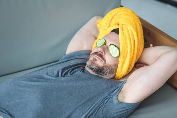 hombre guapo acostado en el sofá con rodajas de pepino en los ojos y toalla amarilla en la cabeza. concepto de autocuidado masculino, cuidado de la piel, relajación en casa. - male towel men human face fotografías e imágenes de stock