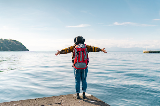 A young female traveller is looking at the sea while outstretching her arms on her vacation.