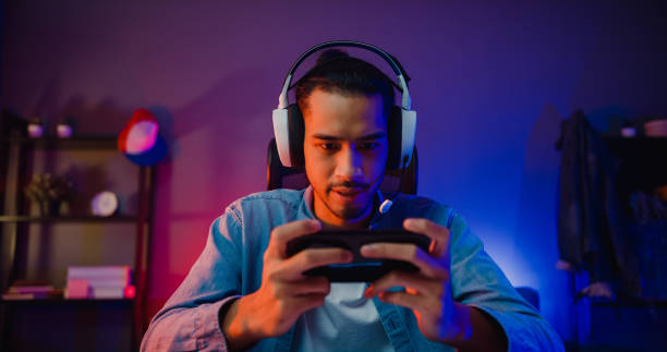 Happy asia man gamer wear headphone competition play video game online with smartphone colorful neon light in living room at night modern house. stock photo