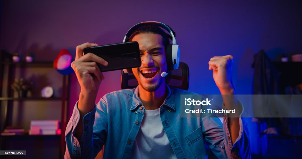 Happy asia man gamer wear headphone competition play video game online with smartphone colorful neon light in living room at night modern house. Happy asia man gamer wear headphone competition play video game online with smartphone colorful neon light in living room at night modern house. Esport streaming game online, Home quarantine activity. Gamer Stock Photo
