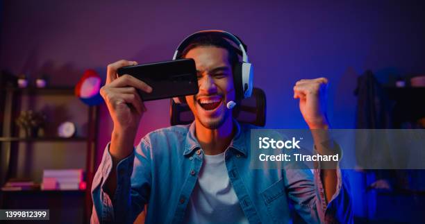 Happy Asia Man Gamer Wear Headphone Competition Play Video Game Online With Smartphone Colorful Neon Light In Living Room At Night Modern House Stock Photo - Download Image Now