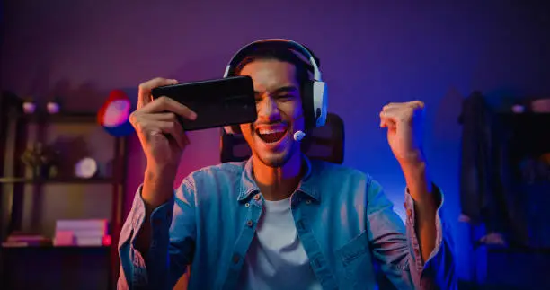 Photo of Happy asia man gamer wear headphone competition play video game online with smartphone colorful neon light in living room at night modern house.