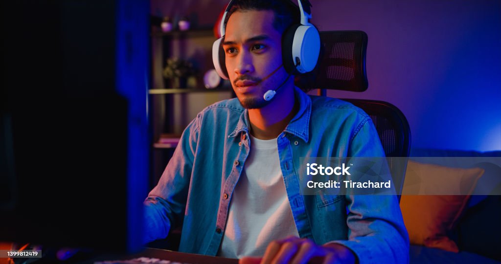 Happy asia guy gamer wear headphone participation play video game colorful neon lights computer in living room at night modern house. Happy asia guy gamer wear headphone participation play video game colorful neon lights computer in living room at night modern house. Esport streaming game online, Home quarantine activity concept. Gamer Stock Photo