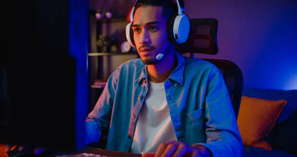 Happy asia guy gamer wear headphone participation play video game colorful neon lights computer in living room at night modern house. stock photo
