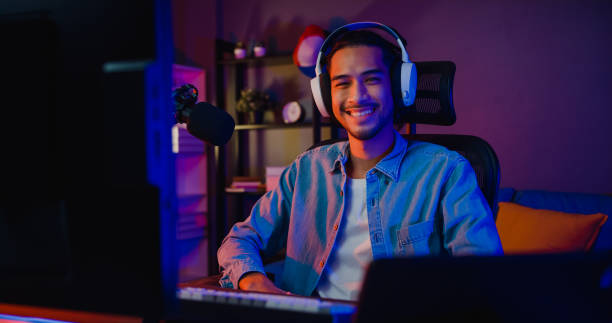 Happy asia guy gamer wear headphone participation play video game colorful neon lights computer and smiling look at camera in living room at night modern house. stock photo