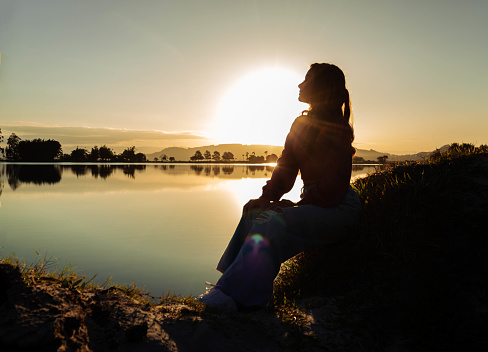 Girl wearing a pink blouse and jeans sitting in front of a lake in a late afternoon with mountains and the sun in the background. Mention travel, relaxation and discovering new places.