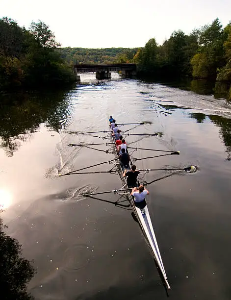 Crew team rowing on river with railroad bridge in background during autumn