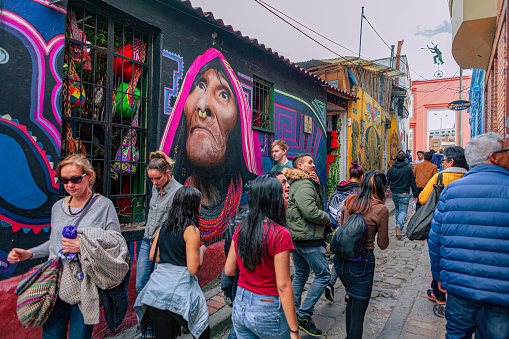 Bogota, Colombia - May 28, 2017: Both Tourists and local Colombian people walk up the narrow Calle del Embudo one of the most colorful streets in the historic La Candelaria district of Bogotá, the Andean capital city of the South American country of Colombia. The street leads to the Chorro de Quevedo, the plaza where it is believed the Spanish Conquistador, Gonzalo Jiménez de Quesada founded the city in 1538. Many street facing walls in this area are painted with either street art or the legends of the pre-Colombian era, in the vibrant colours of Colombia. The altitude at street level is 8,660 feet above mean sea level. Photo shot on an overcast morning; horizontal format.