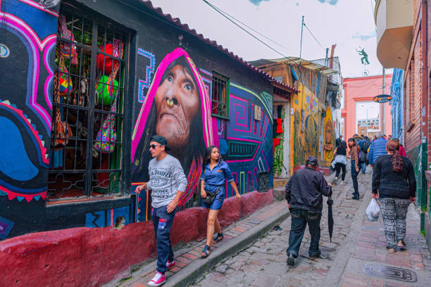 Bogota, Colombia - Tourists And Local Colombians On The Calle del Embudo, In The Historic La Candelaria District Of The Andes Capital City In South America. Bogota, Colombia - May 28, 2017: Both Tourists and local Colombian people walk up the narrow Calle del Embudo one of the most colorful streets in the historic La Candelaria district of Bogotá, the Andean capital city of the South American country of Colombia. The street leads to the Chorro de Quevedo, the plaza where it is believed the Spanish Conquistador, Gonzalo Jiménez de Quesada founded the city in 1538. Many street facing walls in this area are painted with either street art or the legends of the pre-Colombian era, in the vibrant colours of Colombia. The altitude at street level is 8,660 feet above mean sea level. Photo shot on an overcast morning; horizontal format. calle del embudo stock pictures, royalty-free photos & images