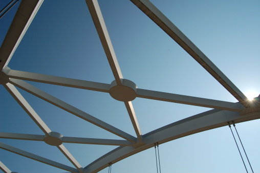 Detail of trusses and cables of a suspension bridge against a blue sky, highlighted by the sun.
