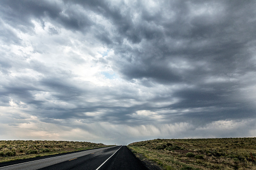 Low angle shot of a summer extreme weather dark rain storm dramatic sky cloudscape above an uphill diminishing perspective asphalt highway road splitting an empty grassy prairie wilderness area somewhere in late afternoon Wyoming, during a western USA road trip.