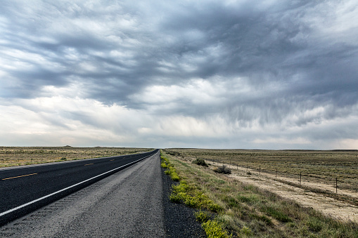 Wide shot of an angry rain storm extreme weather dramatic sky cloudscape high above a long, straight, diminishing perspective dark asphalt highway road splitting the empty, flat, grassy prairie wilderness on a late afternoon in Wyoming, during summer a western USA road trip.
