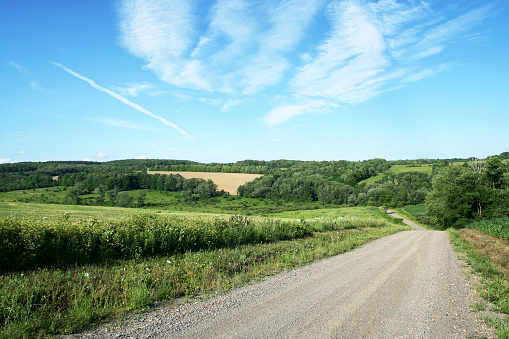 A calm, idyllic, central New York State scene looking down a rolling hill on a tranquil rural country dirt and gravel road winding through quiet and peaceful agricultural fields, lush meadows and forested areas.