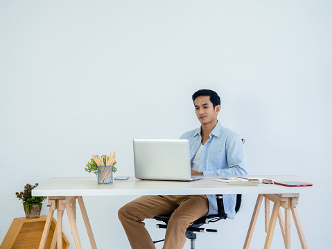 Handsome young Asian man in denim shirt using laptop on table on white clean space wall background in home office. Learning, working, startup with small business, stock, financial, investment concept.